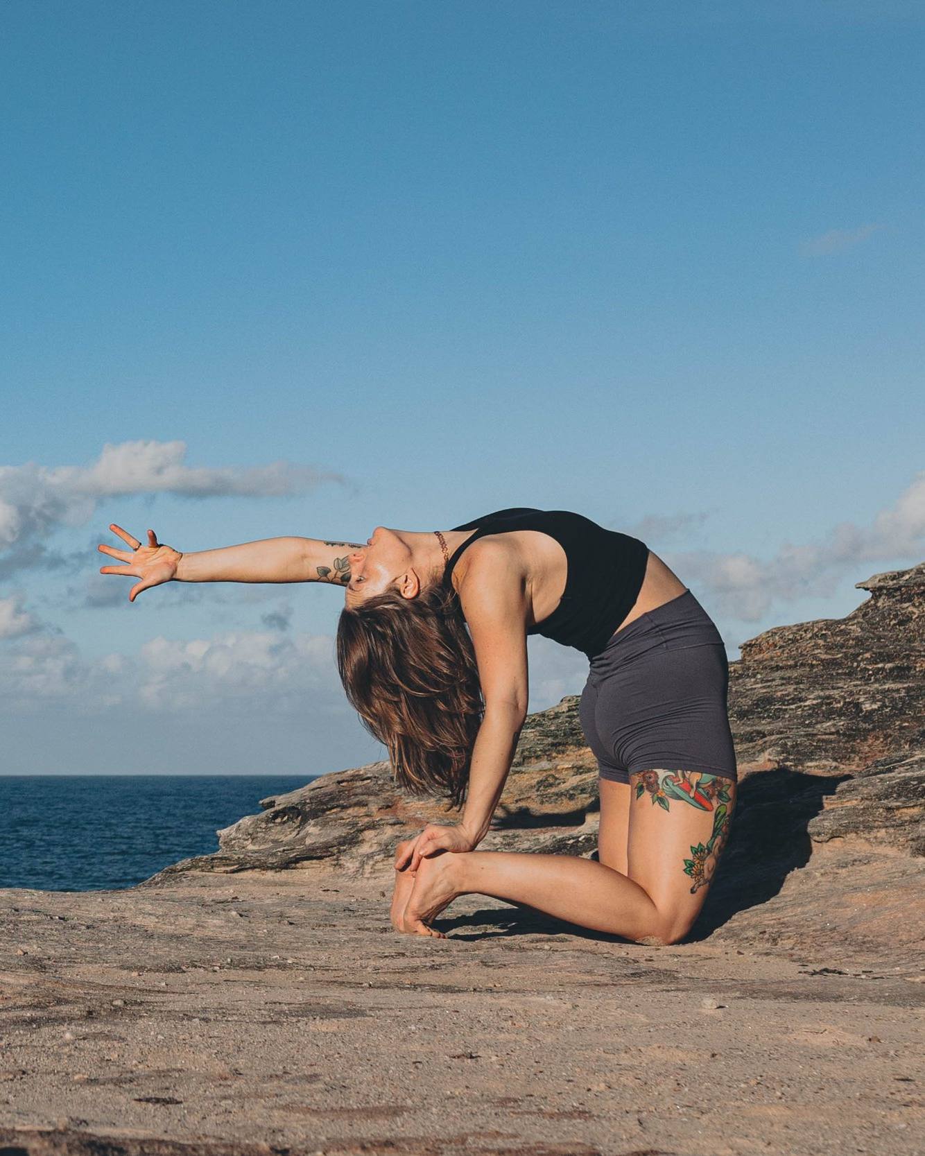 tess kneels on a sandstone ledge in camel pose, one arm reaching back toward the ocean
