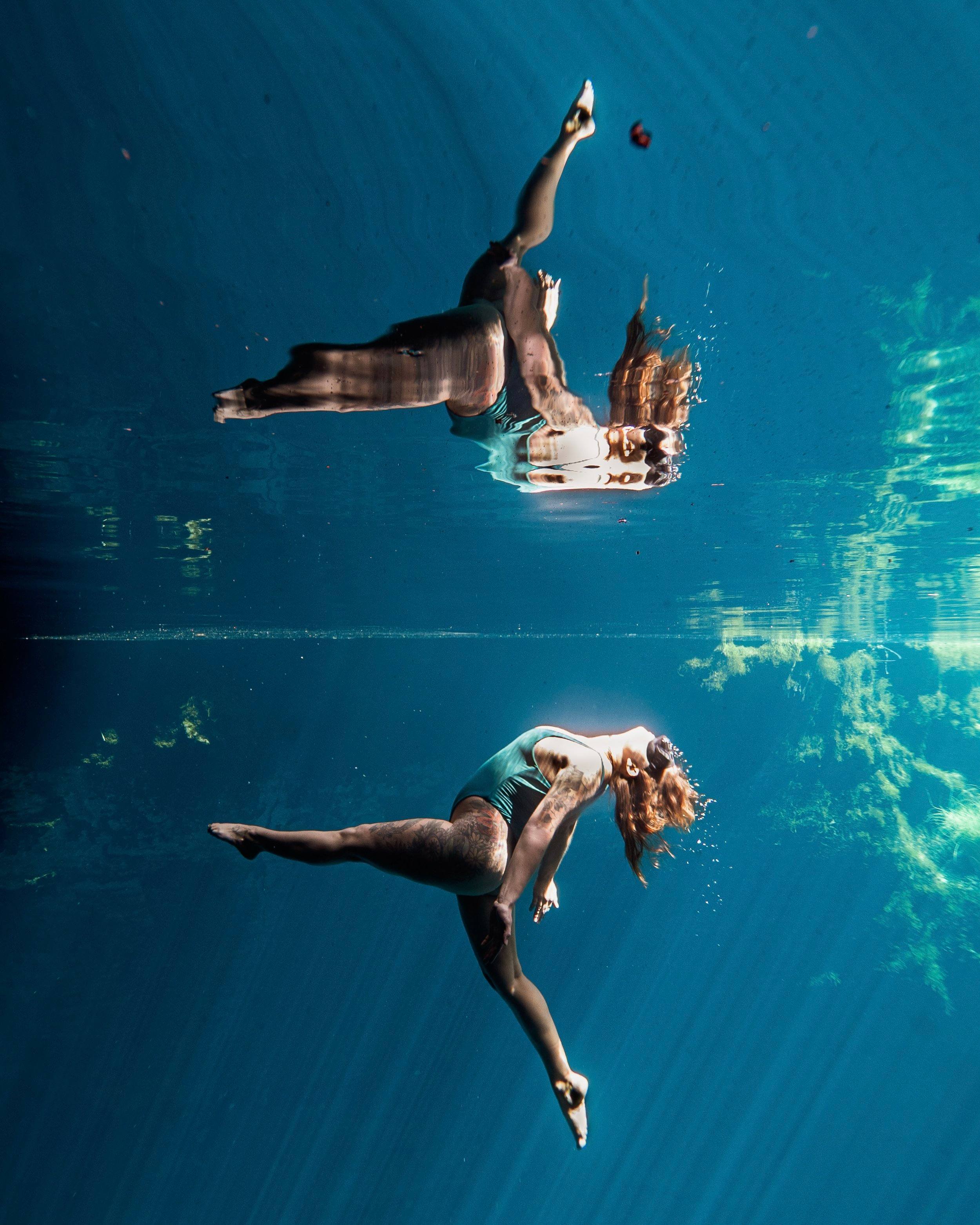 tess floats underwater in a teal swimsuit, her reflection mirrored on the surface above
