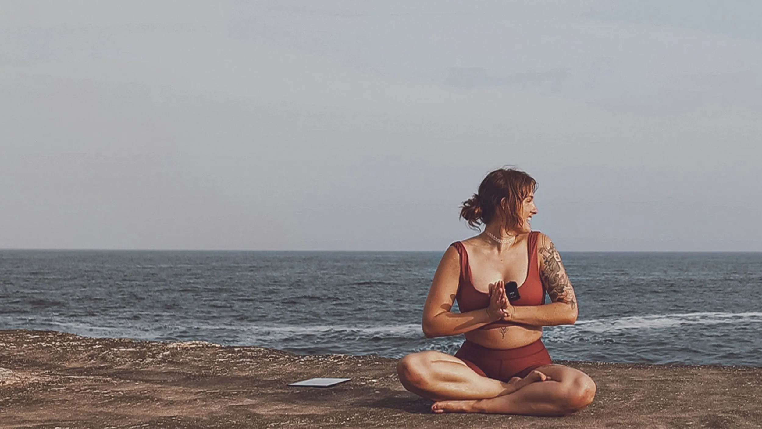 tess sits cross-legged on a rock shelf by the sea, hands in prayer behind her back, laughing